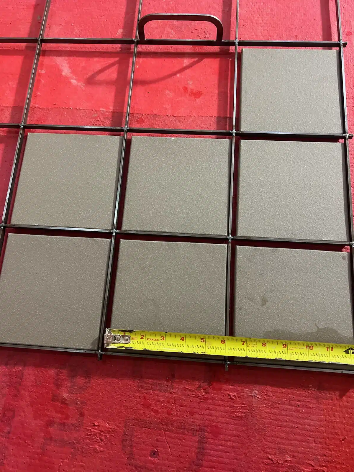 Metal grid frame with textured concrete tiles, measuring approximately 10 inches in width, placed on a red surface. Ideal for construction or tiling projects, showcasing durable material and precise dimensions.
