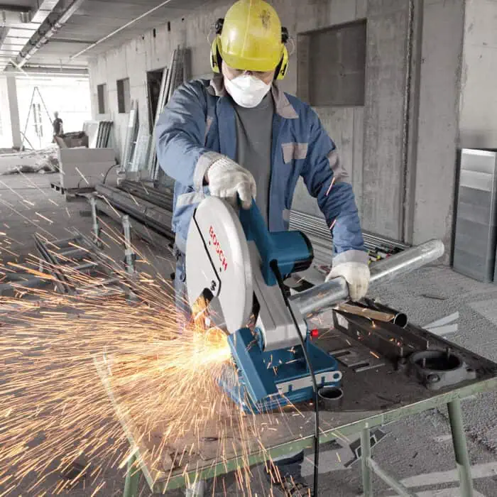 Cutting metal pipe with profissional Bosch circular saw at construction site, sparks flying, worker wearing PPE, safety helmet, gloves, industrial renovation, construction safety, metalworking, building project, construction industry relevance.