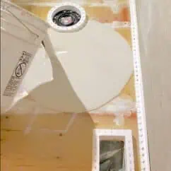 Rough-in plumbing installation for bathroom toilet with subfloor preparation, showcasing drain flange, framed access panel, and wooden subfloor under construction, close-up view of bathroom renovation process.