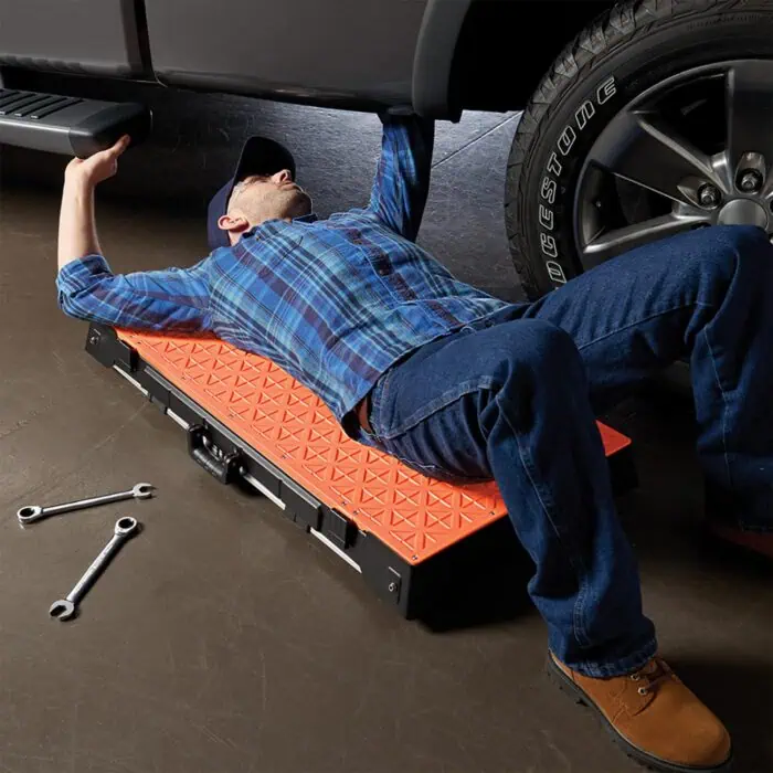 Wheel alignment technician performing vehicle service with orange creeper on garage floor.