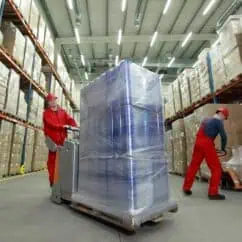 Pallet jack operators moving large wrapped pallets in a high-ceiling warehouse with shelves filled with cardboard boxes, showcasing logistics and inventory management equipment.
