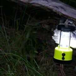 Bright yellow portable lantern hanging near logs and greenery, featuring LED light for outdoor illumination, ideal for camping and survival gear.
